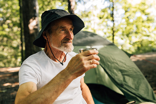 Senior male in hat holding metallic cup while sitting in the forest against tent