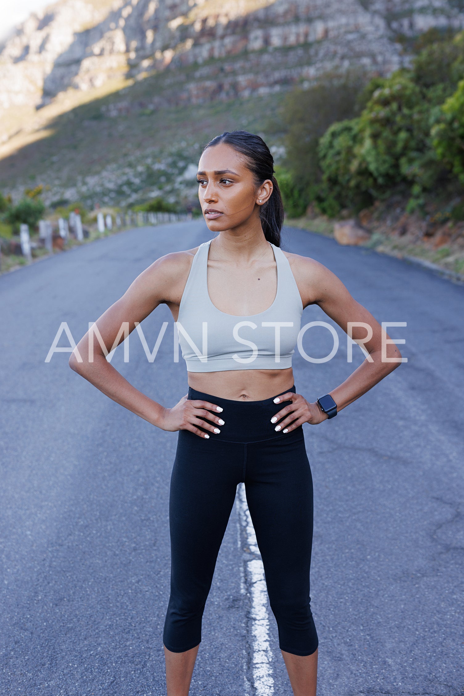 Young confident slim female with hands on her hips standing outdoors on an abandoned road and looking away. Woman jogger relaxing during outdoor workout.