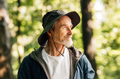 Side view of senior male in hat standing in forest. Portrait of a confident mature tourist posing outdoors.