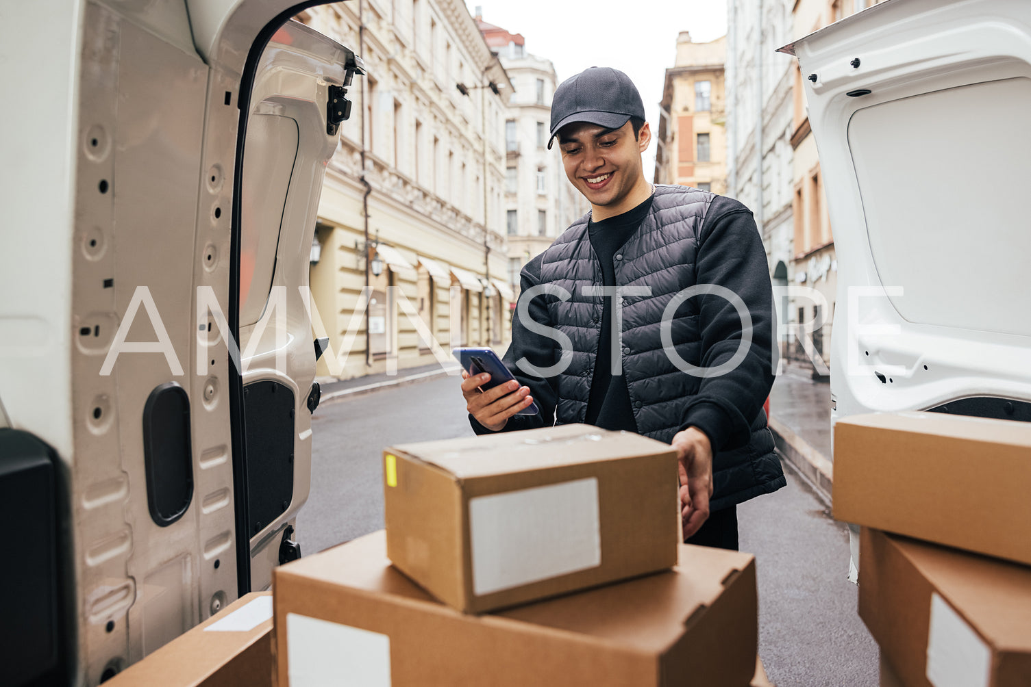 Smiling courier in uniform standing at car trunk checking mobile