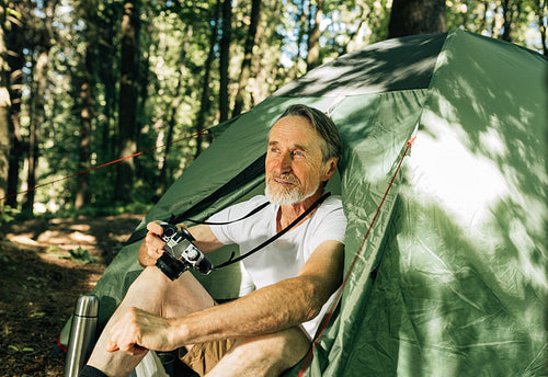 Senior tourist looking away while sitting in tent. Mature male with a film camera enjoying his forest camping.