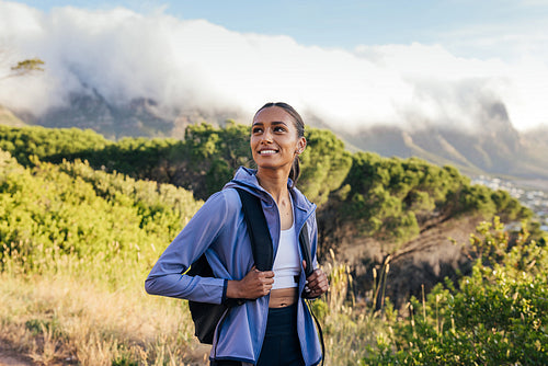 Smiling woman with backpack and sportswear enjoying hiking at sunset. Cheerful woman looking away while walking outdoors in a natural park.