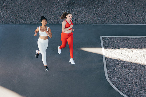 High angle of two females running together. Two women with different body types jog outdoors wearing sportswear with different colors.