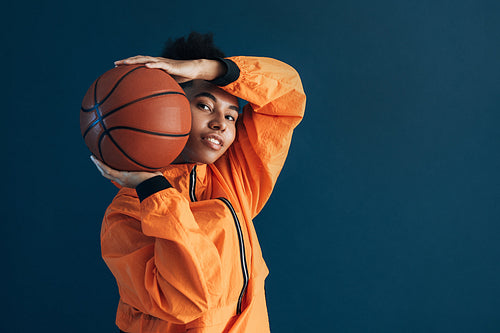 Portrait of a young basketball player posing with ball against blue backdrop and looking at camera