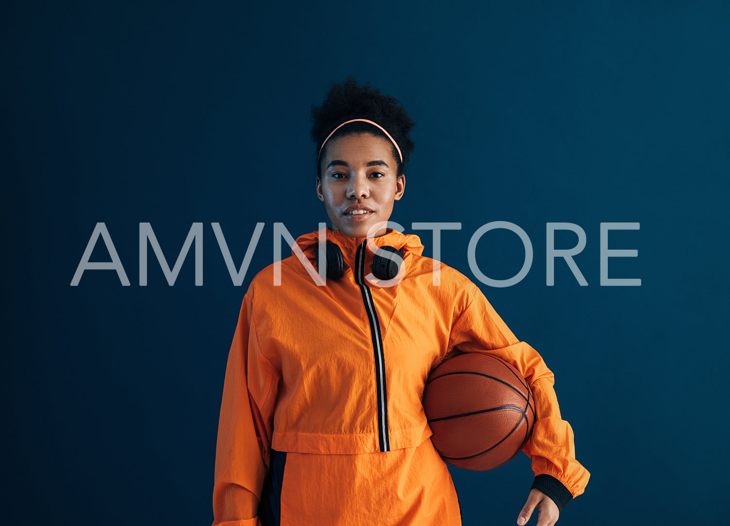 Portrait of a young basketball woman looking straight of a camera wearing orange sportswear. Confident sportswoman with a basketball over blue backdrop.