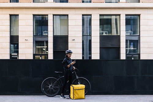 Side view of delivery woman holding a mobile phone, wearing a cycling helmet. Young female standing in the city with bicycle and thermal backpack.