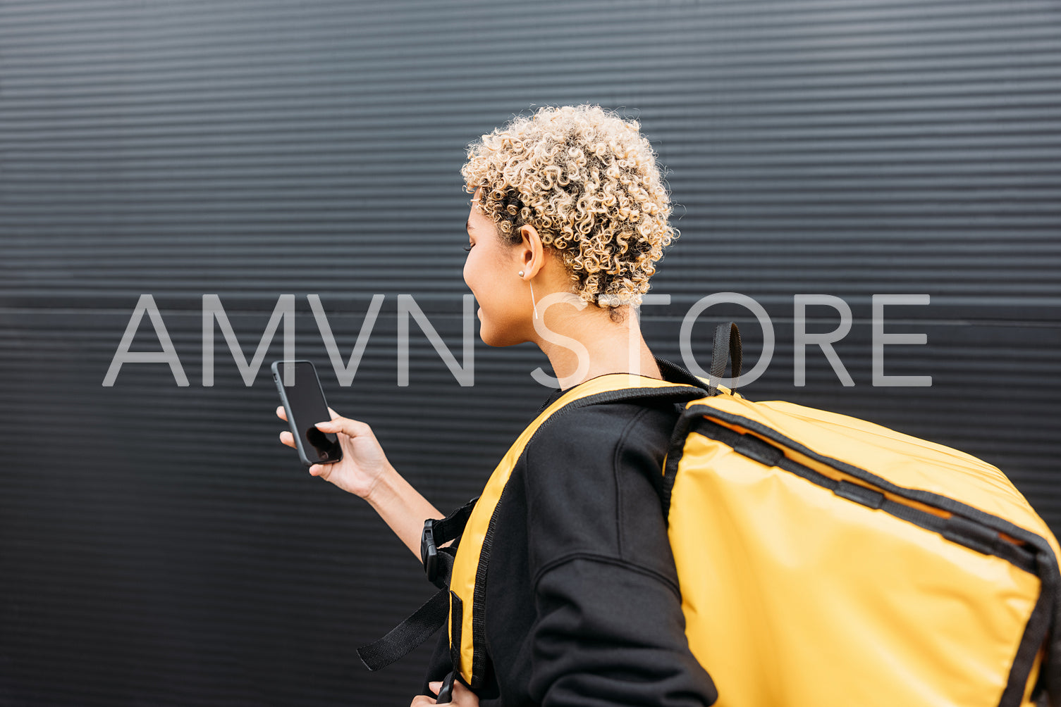 Side view of confident delivery woman with bag standing on a sidewalk holding a smartphone