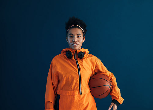 Portrait of a young basketball woman looking straight of a camera wearing orange sportswear. Confident sportswoman with a basketball over blue backdrop.