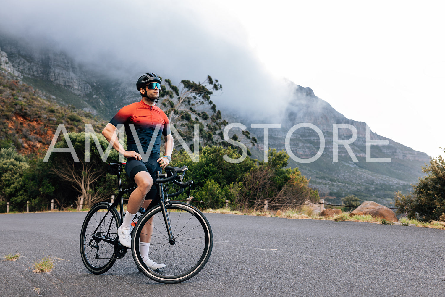 Young male cyclist relaxing after training with hands on a hips. Professional athlete with road bike enjoying the view while standing on a road in wild terrain.