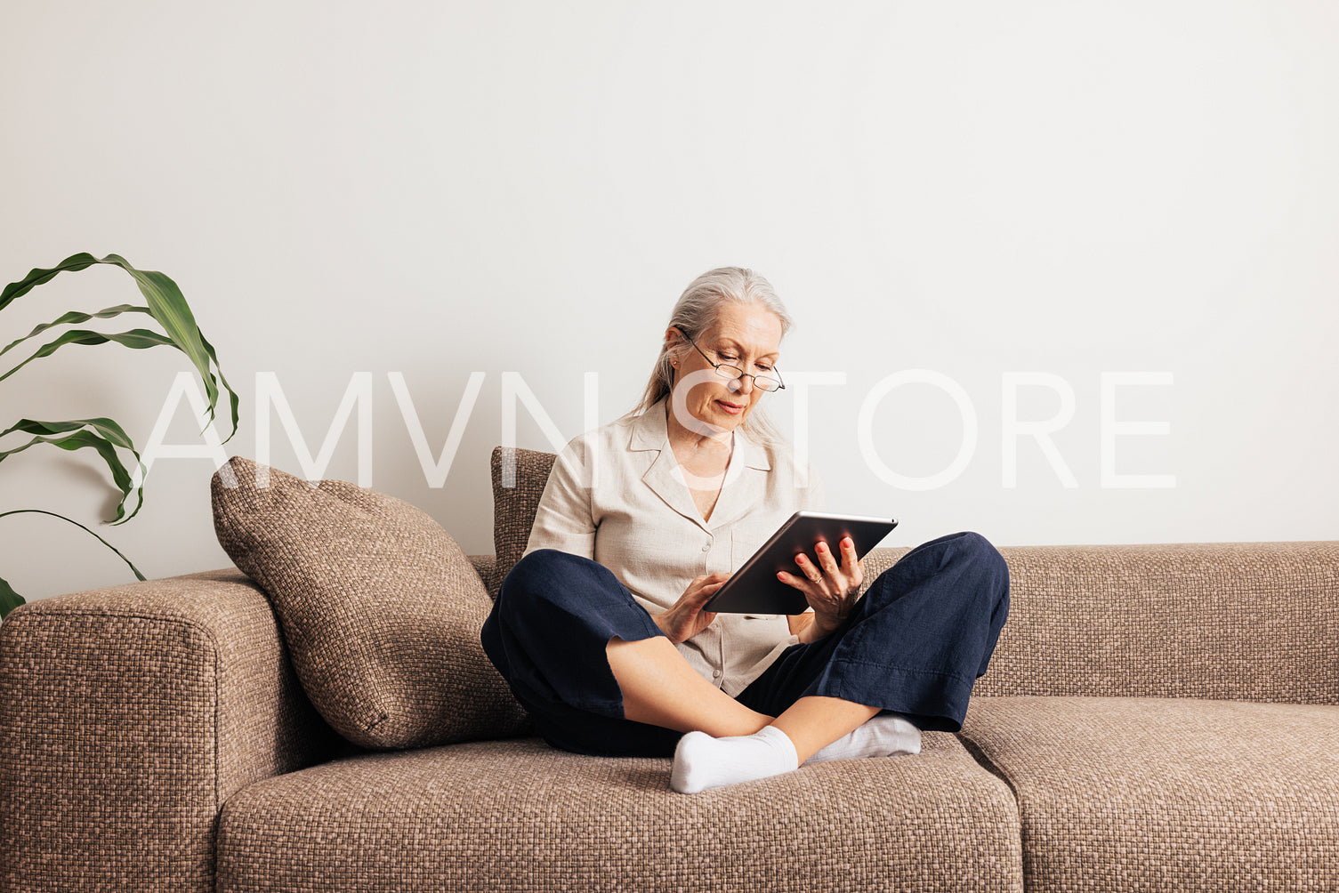 Senior woman sitting on a sofa with crossed legs holding digital tablet. Aged female reading from a portable computer at home.