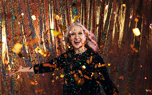 Stylish senior woman in black dress throwing confetti against a