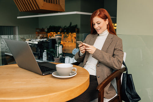 Smiling businesswoman with a smartphone in cafe. Female with ginger hair in formal wear sitting at a table.