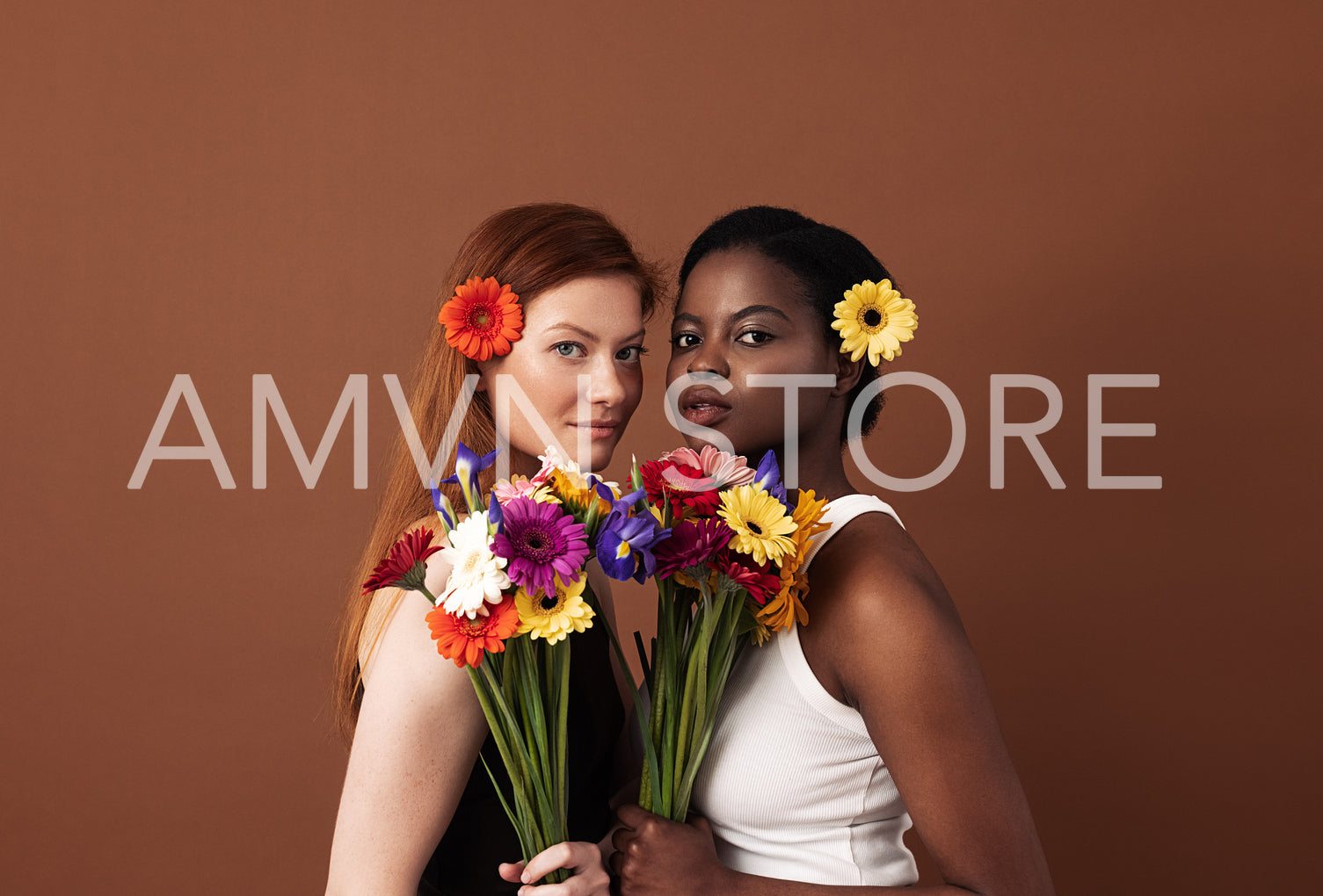 Two women of different races with flowers in their hairs holding bouquets. Caucasian and African American females looking at camera in a studio.