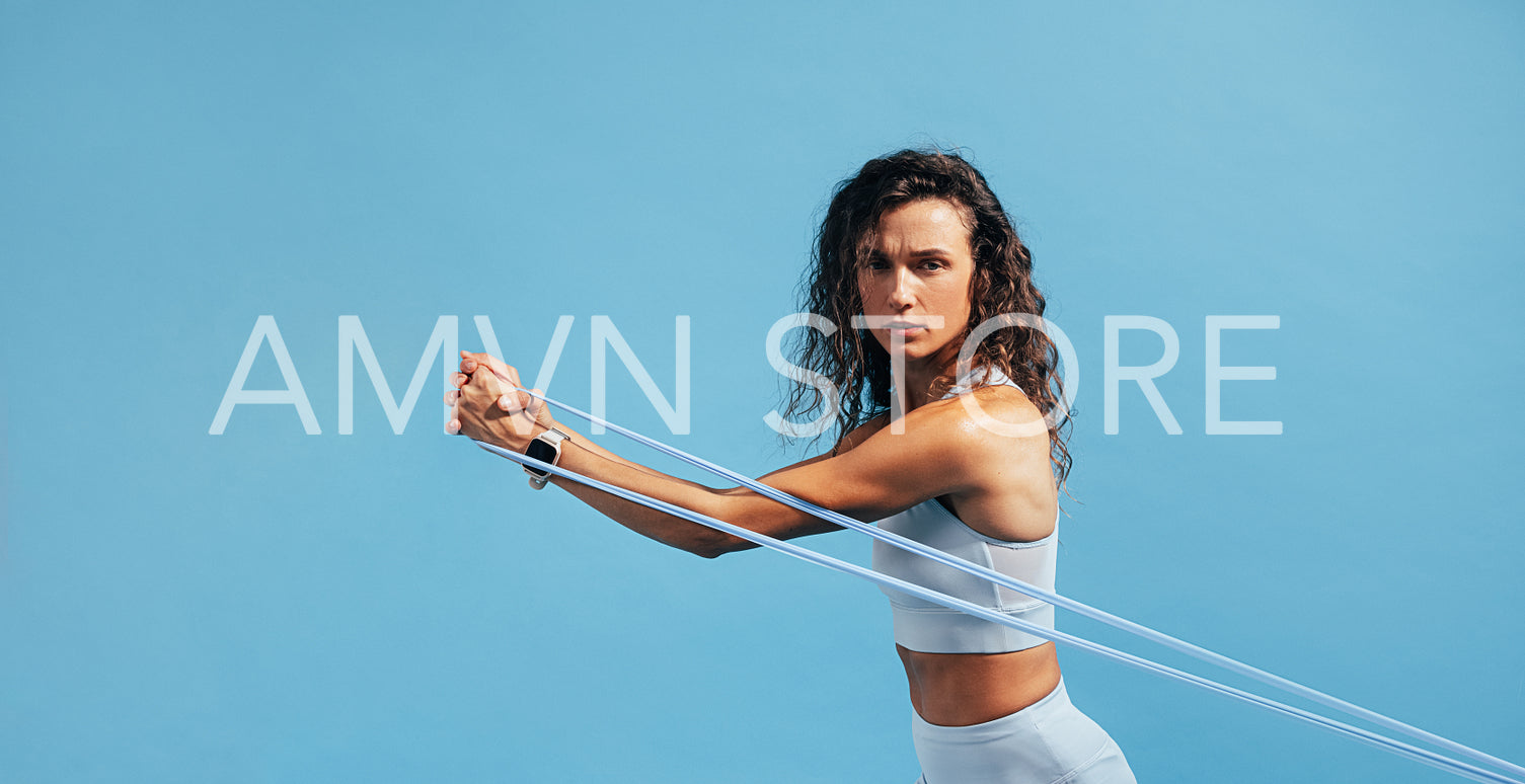 Young muscular woman exercising with a resistance band against blue background