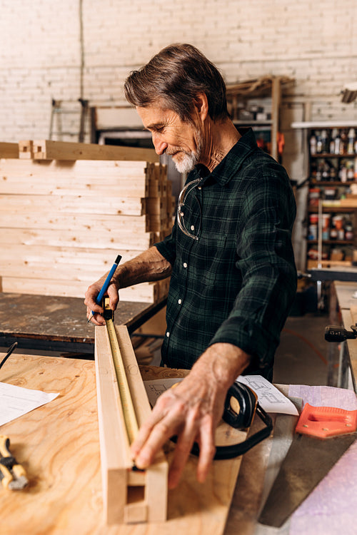 Side view of senior carpenter doing markings on a wooden bar with using a measuring tape