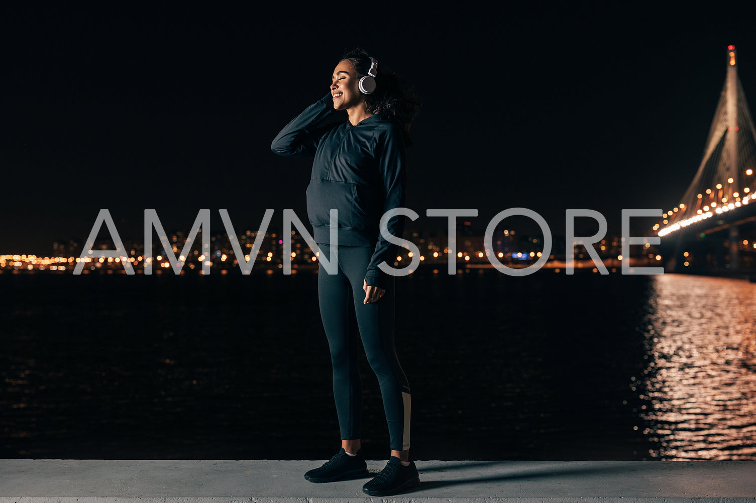 Young female in sport clothes standing on embankment listening to music against night city lights