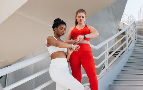 Two plus-size females standing together checking their smartwatches. Full-figured women in fitness attire relax after workout checking their pulse on smartwatches.