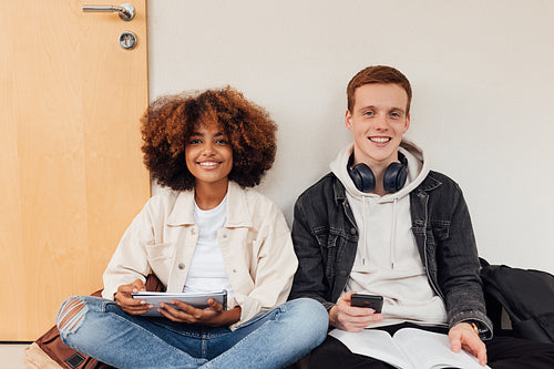 Two smiling classmates looking at camera. Cheerful students sitting together at wall with books.