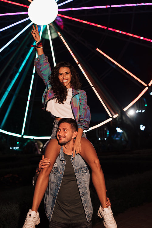 Smiling female sitting on the shoulders of her boyfriend and raising her hand in an amusement park at night