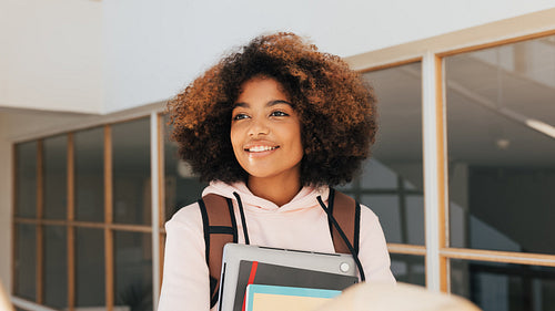 Portrait of a smiling girl with curly hair holding laptop and books while going up on staircase