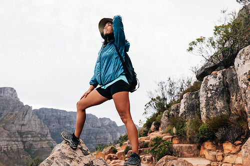 Smiling woman hiker in sports clothes holding a hat on her head while looking up and enjoying the view