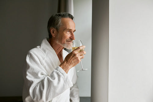 Senior man in bathrobe smell wine looking at window in hotel room