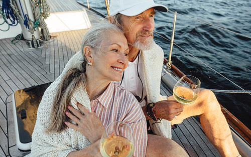 Senior couple wrapped with plaid and sitting on a yacht deck with bocals of wine. Two loving mature people sitting together on a sailboat and looking away.