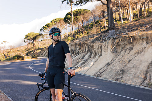 Portrait of a professional cyclist in a helmet looking away while standing on a road. Athlete in black sportswear with road bike relaxing on a country road.