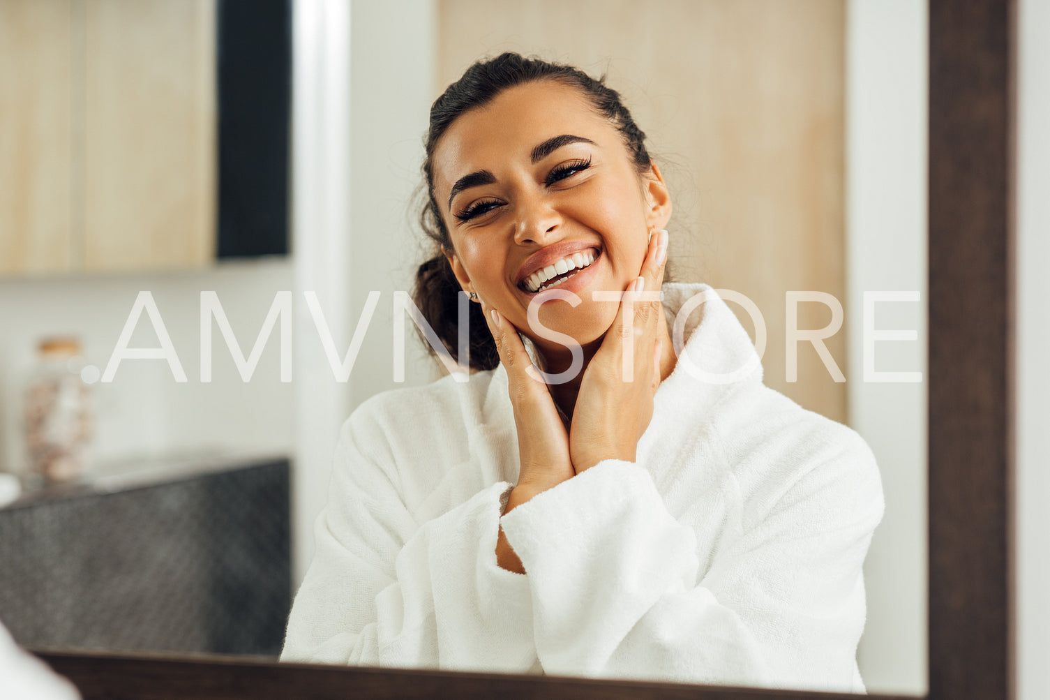 Happy woman in white bathrobe touching her cheeks. Young female looking in her reflection in bathroom mirror.	