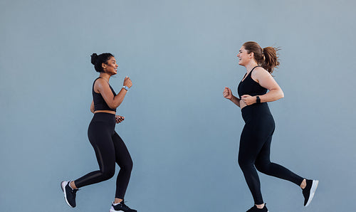 Sportswoman running in front of her friend and motivating to run. Two jogging friends smiling while running at grey wall.