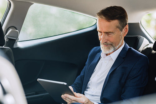 Senior entrepreneur typing on a digital tablet while sitting on the backseat in taxi