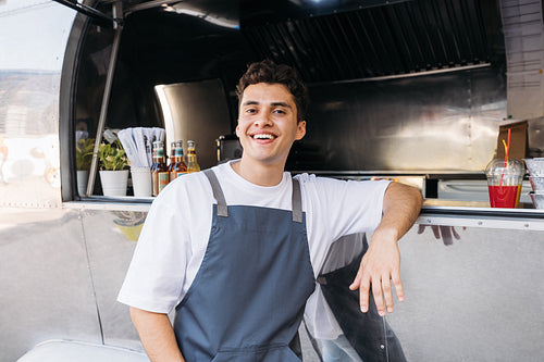 Portrait of a happy food truck owner. Middle eastern guy in apron leaning on food truck looking at camera.