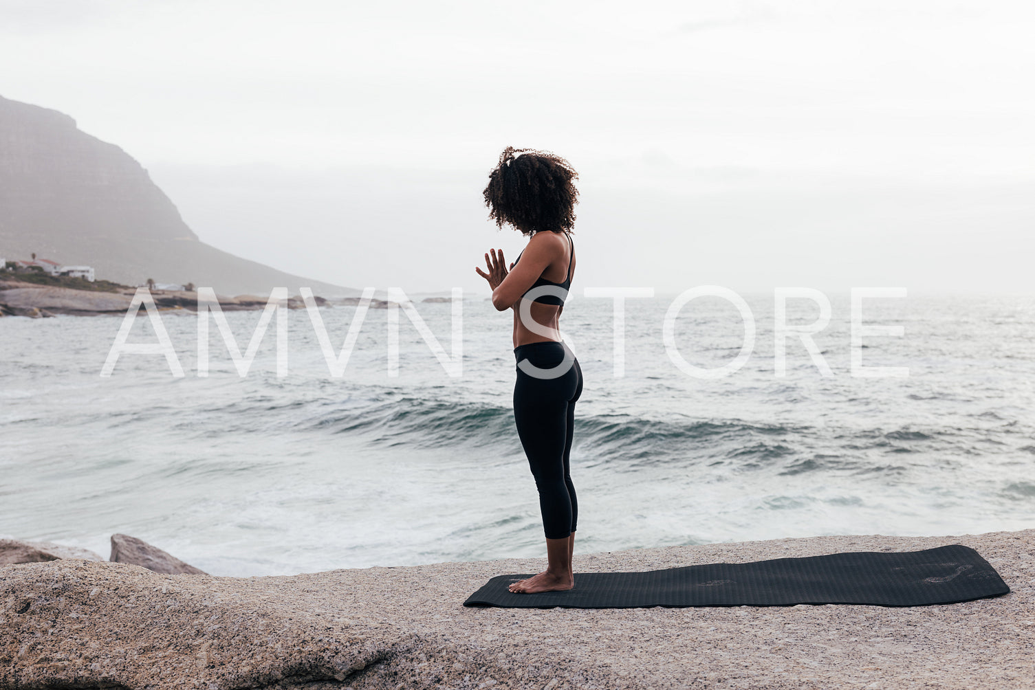 Side view of a slim woman meditating while standing on a mat against an ocean at evening