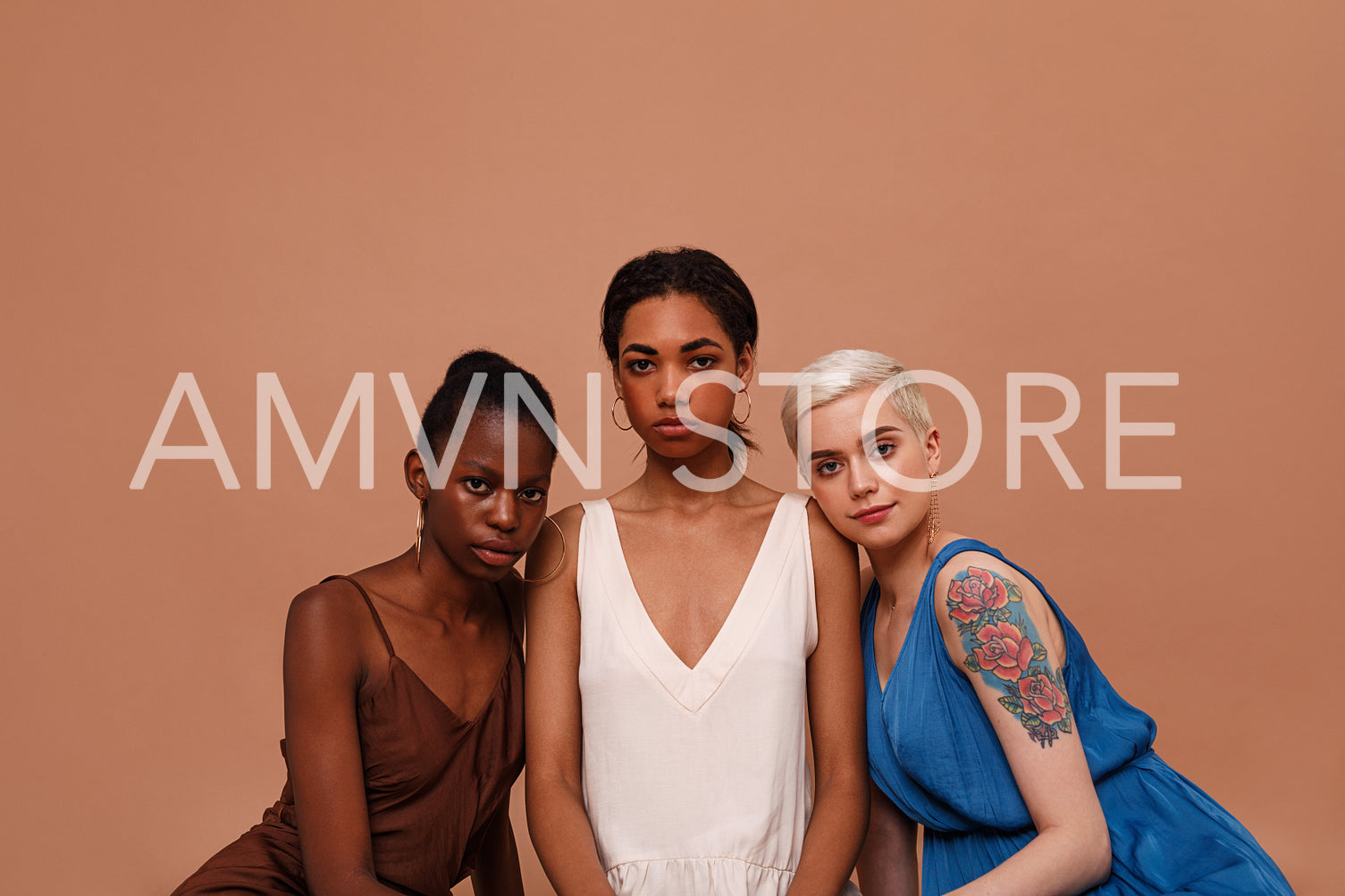 Three beautiful women with different skin color looking at camera. Young females in dresses sitting on brown backdrop.