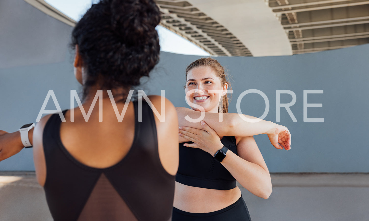 Plus size female smiling and looking at her fitness buddy while stretching her arm. Two women in black fitness attire warming up hands before a workout.