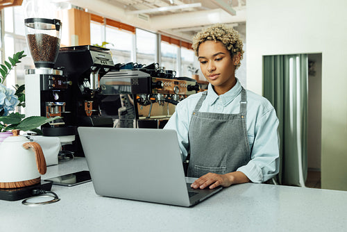 Young female barista working on laptop in a coffee shop. Woman with short hair in an apron at her small local cafe.