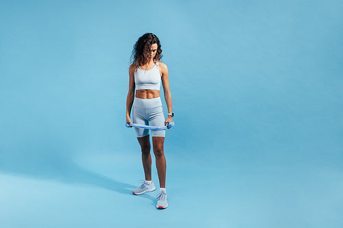 Young slim woman in fitness attire holding a resistance band and looking down on blue background