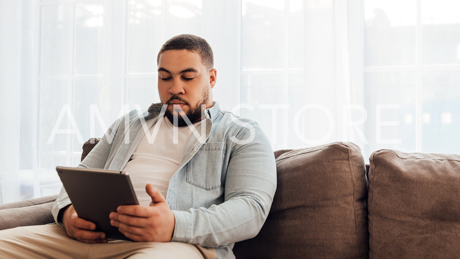 Young man in casual clothes sitting on sofa at home and using digital tablet	