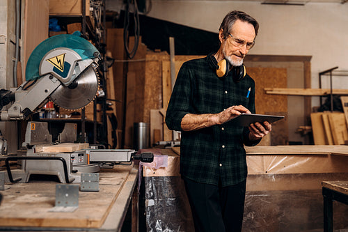 Mature carpenter holds a digital tablet. Male worker standing at workbench in carpentry.