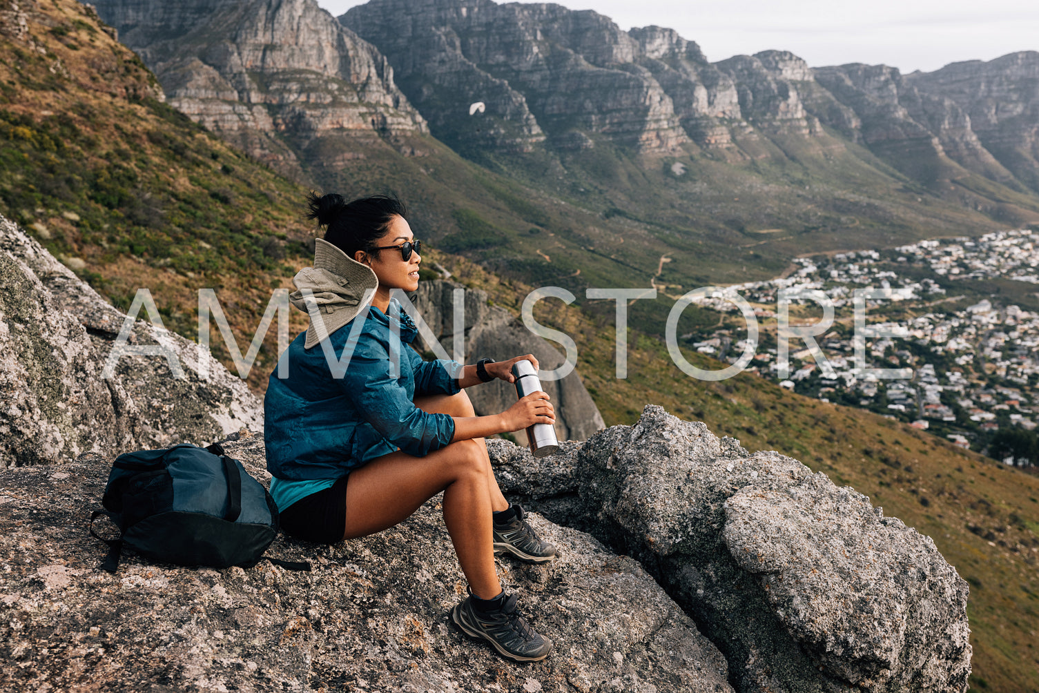 Side view of woman sitting on a rock holding thermos. Female hiker relaxing while sitting on top of a mountain.