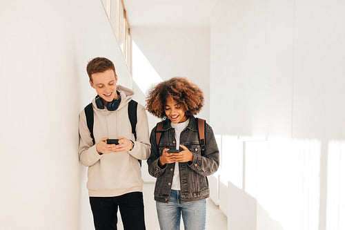 Laughing classmates walking together in corridor holds smartphones