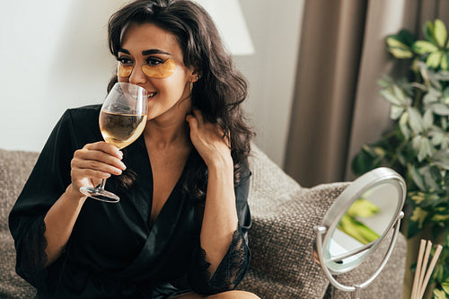 Brunette woman with eye patches drinking wine. Young female in satin robe relaxing at home on couch.