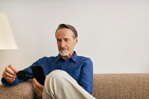 Mature man in casuals buying by credit card with a smartphone on a sofa