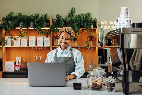 Young smiling woman in an apron typing on a laptop. Female working as a barista standing at the counter with a laptop.