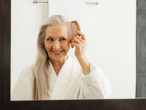 Woman holding a wood comb in the bathroom. Aged female combing her hair while looking at a mirror.