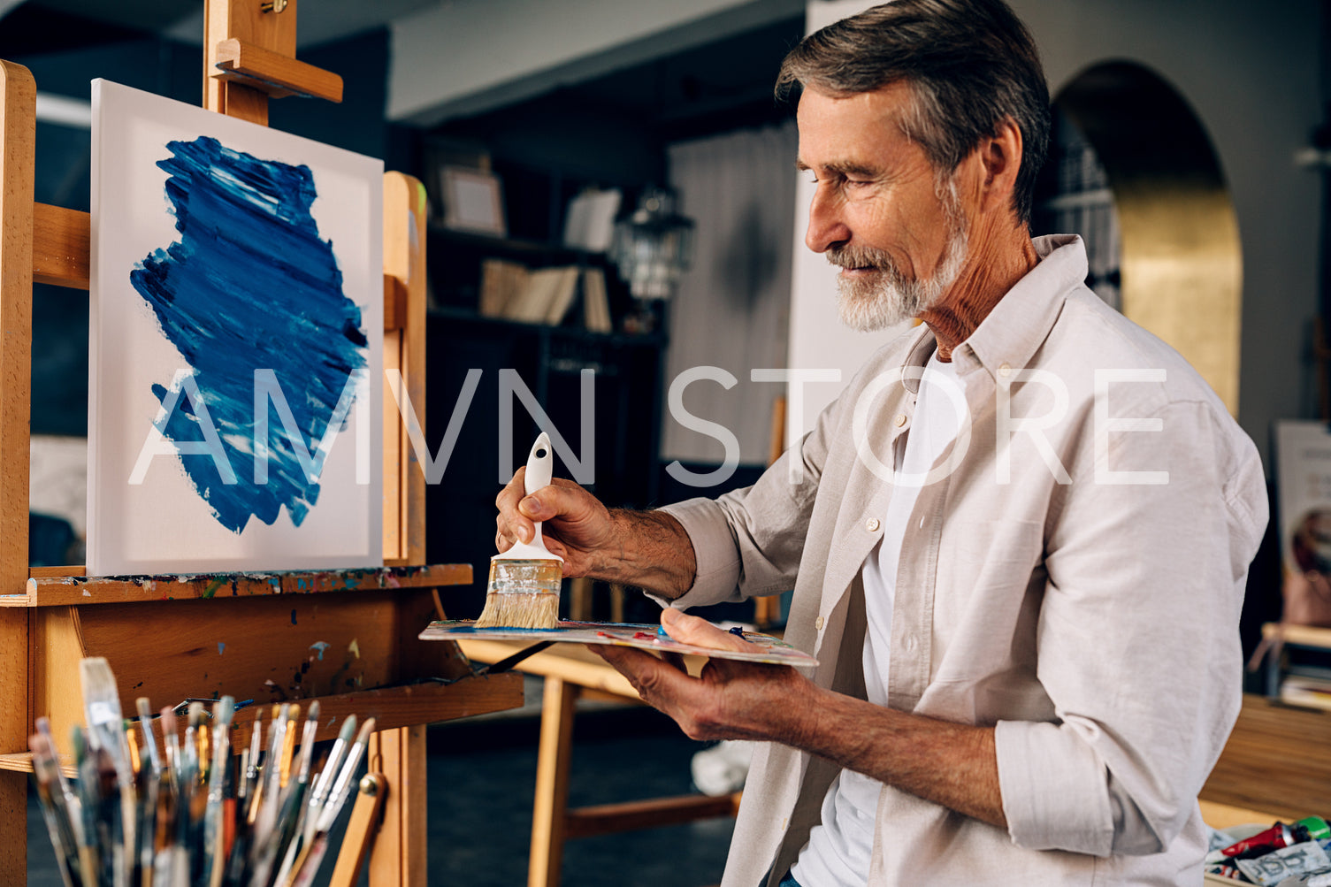 Artist in the studio. Concentrated bearded man sitting in front of easel holding a paintbrush and mixing colors on palette.	