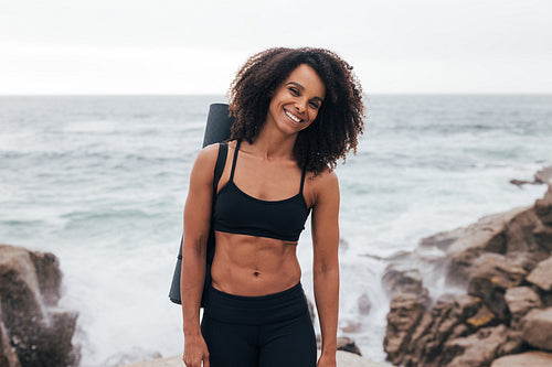 Portrait of a young sportswoman with mat looking at camera while standing by coast