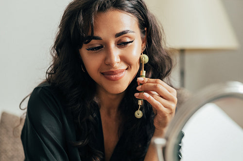 Close up of woman massaging face with jade roller while sitting in front of a mirror