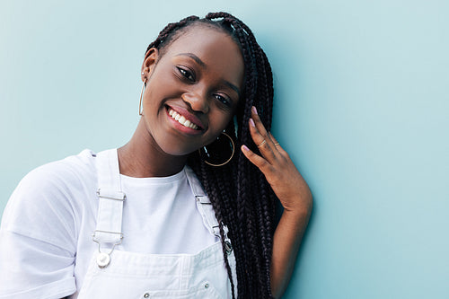 Close-up portrait of a young woman with braids. Smiling female in white clothes. Positive woman leaning blue wall and adjusting her braids while looking at camera.