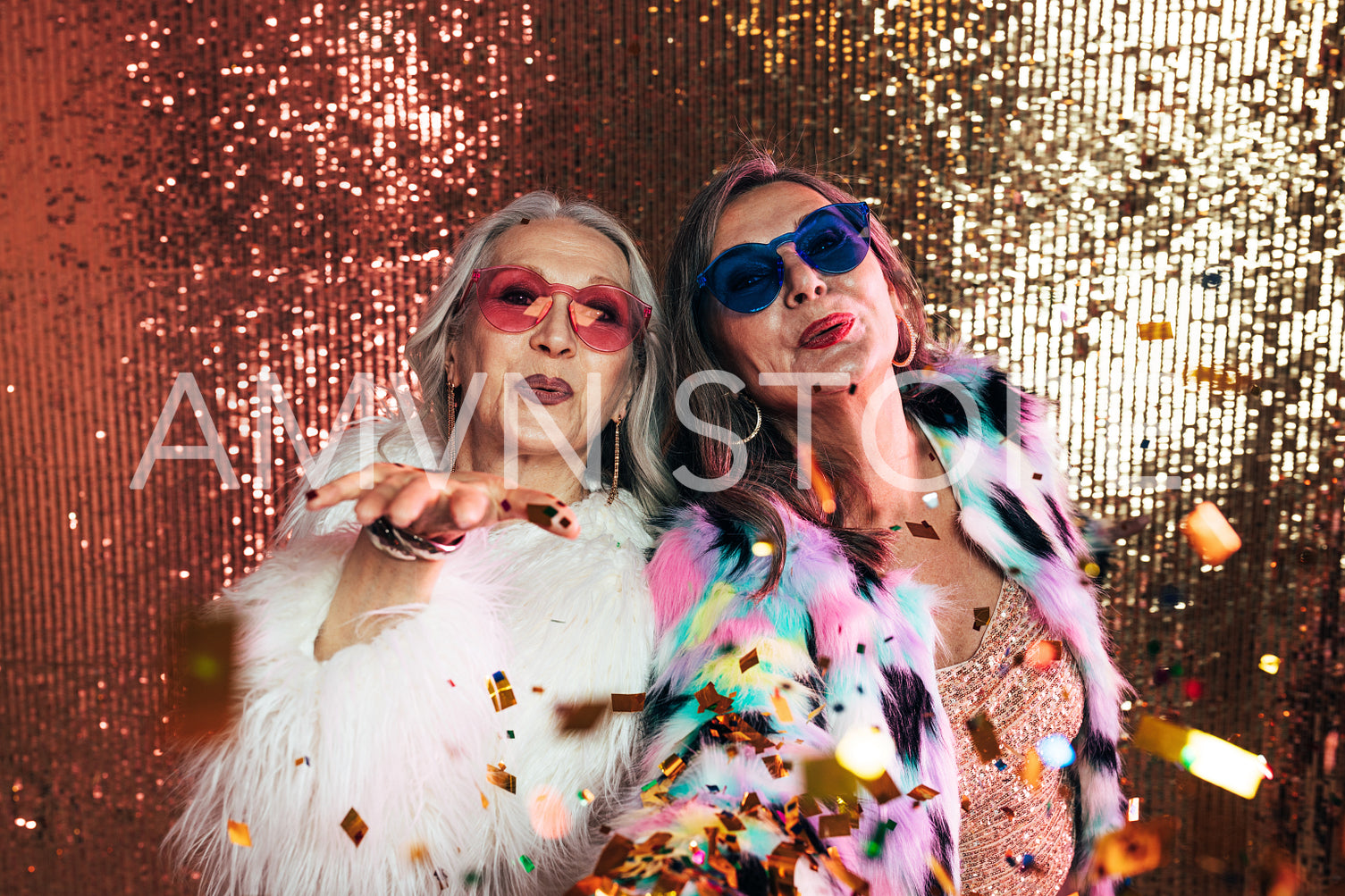 Two senior women in eyeglasses wearing fur coats blowing confett